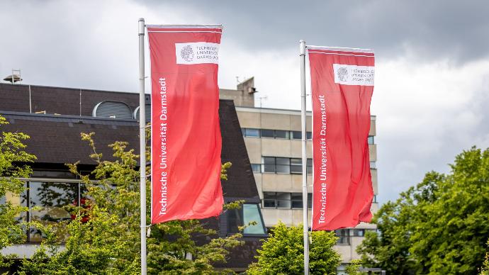 Two red flags with the logo and name of 'Technische Universität Darmstadt' wave in the wind in front of a modern university building with large windows and a dark sloped roof. Green trees partially obscure the building, and the sky is cloudy.