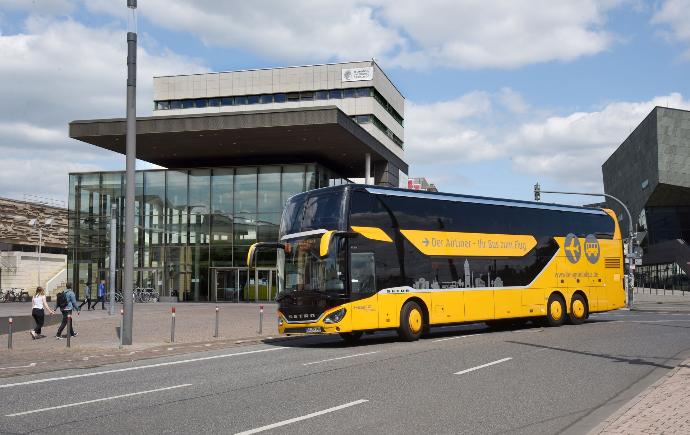 A yellow double-decker long-distance bus with the logo of 'Der Airport – My Shuttle to Flight' is driving on a road in front of a the TU Darmstadt main entrance, a modern glass building with multiple levels. Several pedestrians and cyclists are visible near the entrance. The sky is partly cloudy, and the surrounding architecture is contemporary with clean lines and large windows.