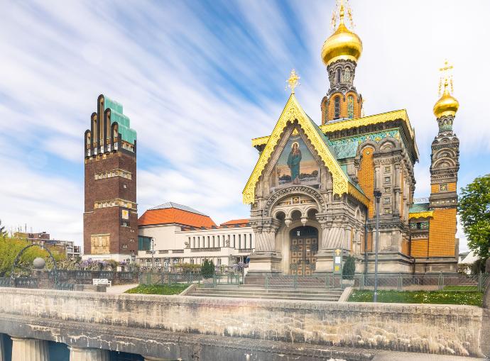 The Wedding Tower of Darmstadt, a tall brick structure with a distinctive stepped roof, stands on the left, accompanied by the Mathildenhöhe Museum with its white facade and red-tiled roof. On the right, the Russian Chapel with its ornate golden domes, richly decorated facade, and intricate mosaics is visible. The sky is filled with streaked clouds, and the foreground features a stone wall and greenery, adding depth to the historic scene.